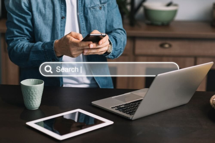 A person sits at a desk with an open laptop, tablet, and cup while using a smartphone. A search bar overlay is displayed across the photo.