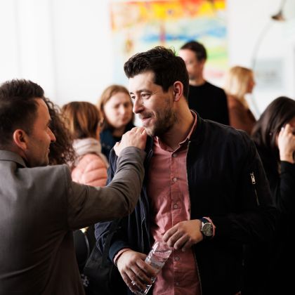 V ospredju sta dva moška obrnjena eden proti drugemu, levi moški ima roko na rami desnega. Oblečena sta poslovno, desni drži v roki plastenko vode. V ozadju je veliko ljudi.