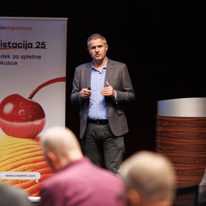 Na sredini je poslovno oblečen moški, ki v roki drži daljinski upravljalnik za diaprojektor. V ospredju se vid nekaj ljudi, ki ga gledajo. V ozadju je pano z napisom "Digistacija 25".