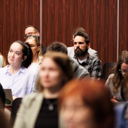 Ljudje, ki sedijo v konferenčni dvorane. prve tri vrste ljudi so motne, jasno je vidna le četrta vrsta ljudi. V ozadju je lesena stenska obloga.