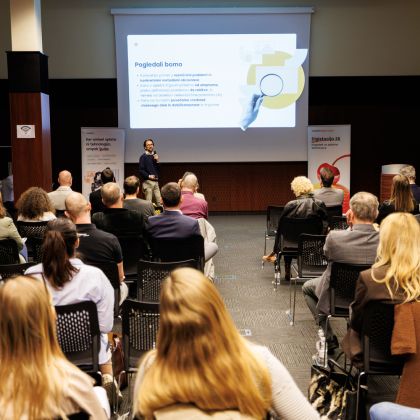 Konferenčna dvorana. Vidnih je šest vrst stolov, na katerih sedijo ljudje in gledajo govorca, ki je viden v ozadju. Na steni zadaj je predstavitev na platnu z naslovom "Pogledali bomo".