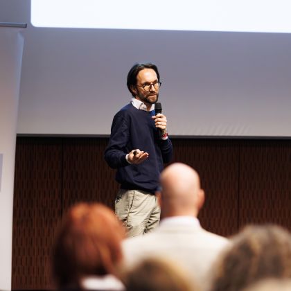 Moški z očali v desni roki drži daljinski upravljalnik za diaprojektor, v levi pa mikrofon. V ozadju je pano z napisom "Smisel spleta ni tehnologija, ampak ljudje". v ospredju se vidi nekaj glav ljudi, ki gledajo govorca.