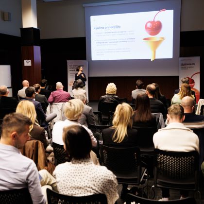 Konferenčna dvorana. Vidnih je šest vrst stolov, na katerih sedijo ljudje in gledajo govorca z mikrofonom, ki je viden v ozadju. Na steni zadaj je predstavitev na platnu z naslovom "Ključna priporočila".