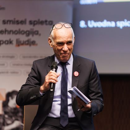 Moški v obleki s kravato sedi na stolu. V desni roki ima mikrofon, v levi pa blokec in kemični svinčnik. Za njim je pano, na katerem piše "Smisel spleta ni tehnologija, ampak ljudje".