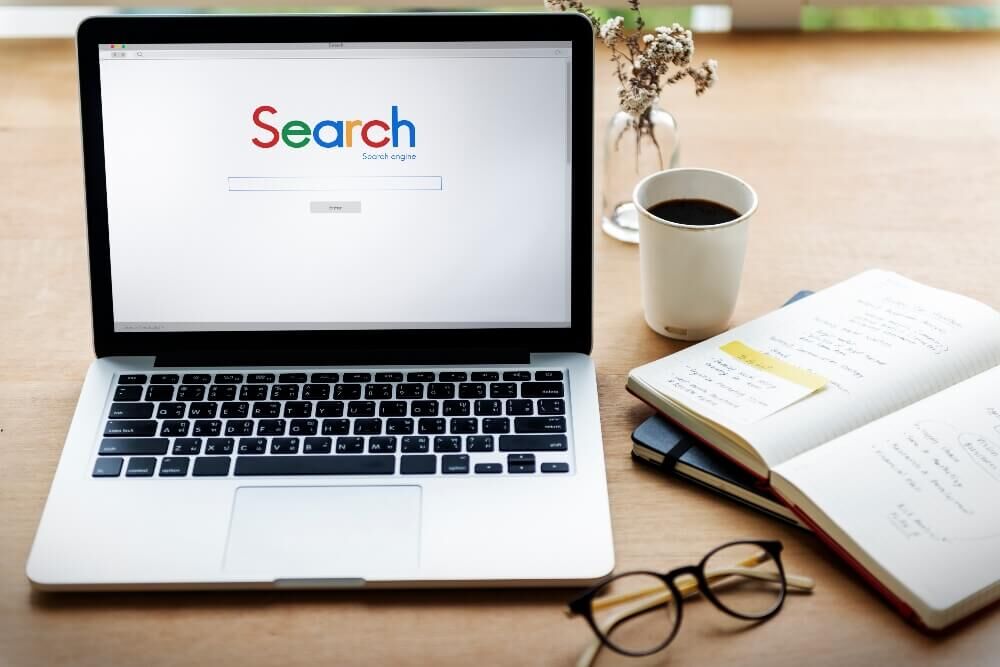 A laptop on a desk shows a search engine on the screen. Next to it are a cup of coffee and an open notebook with notes.